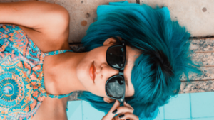 a girl with vivid blue colored hair wearing sunglasses by a pool.